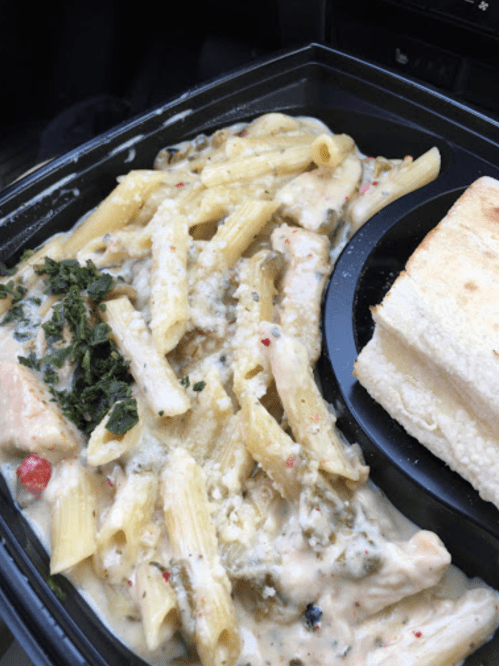 A takeout container with creamy pasta, chicken, vegetables, and a slice of bread.